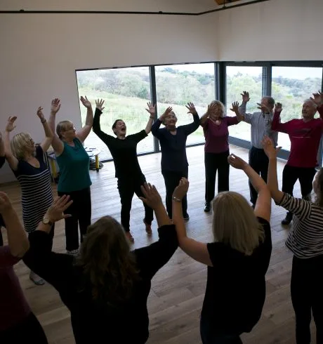 A group of people are standing in a circle with their arms in tehe air. They are in a studio with large picture windows showing countryside outside