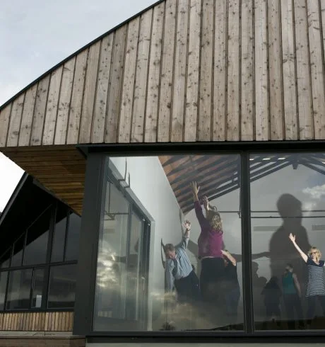 A group of people are taking part in a movement class inside a modern building that references rural steadings , clad in timber with large picture windows on two sides.