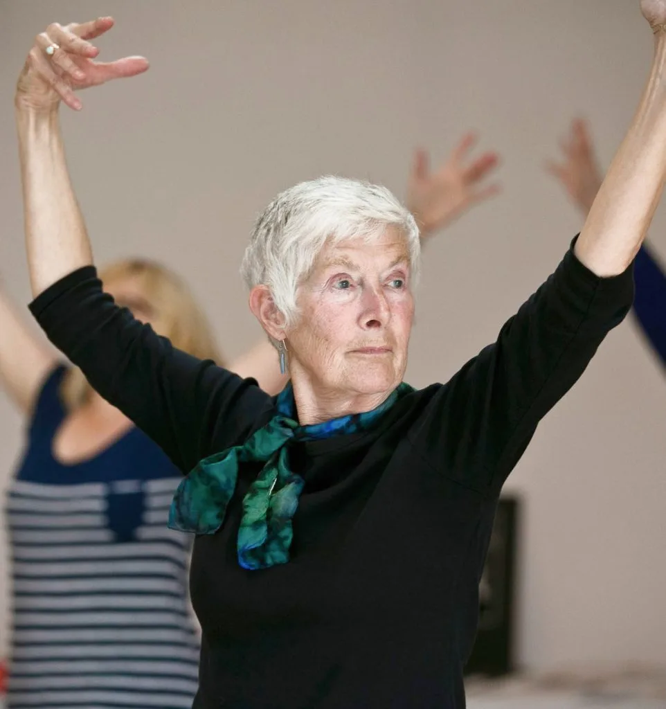 An older woman with short silver hair is holding her arms up in a dance pose