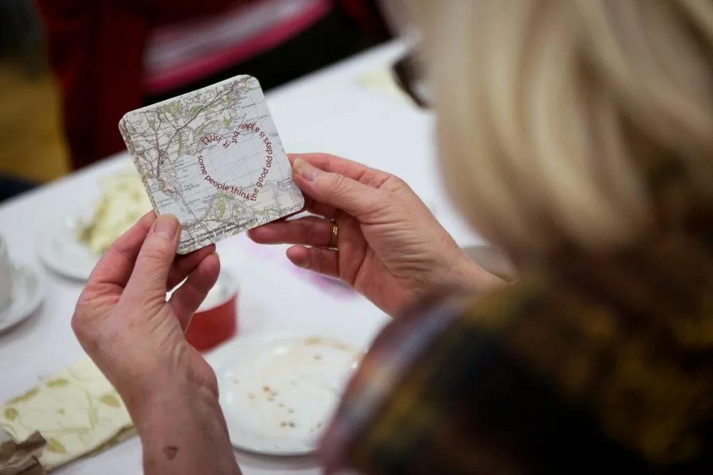 A woman is looking at a coaster made from an old map, which has been over printed by text in the shape of a heart that reads "Some people think the good old days is a joke but it isn't .."