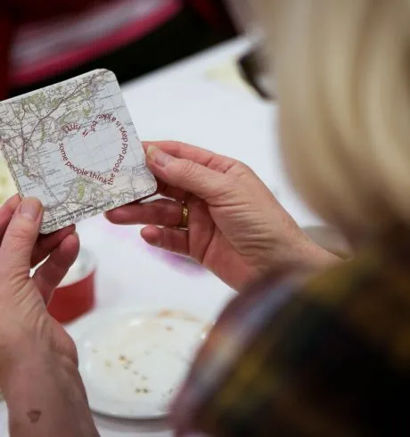 A woman is looking at a coaster made from an old map, which has been over printed by text in the shape of a heart that reads "Some people think the good old days is a joke but it isn't .."