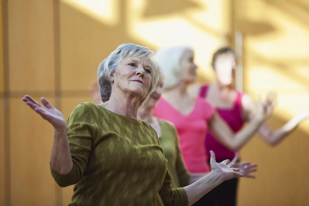 An older woman is performing a dance routine with others. She is leaning back slightly with her arms semi-raised