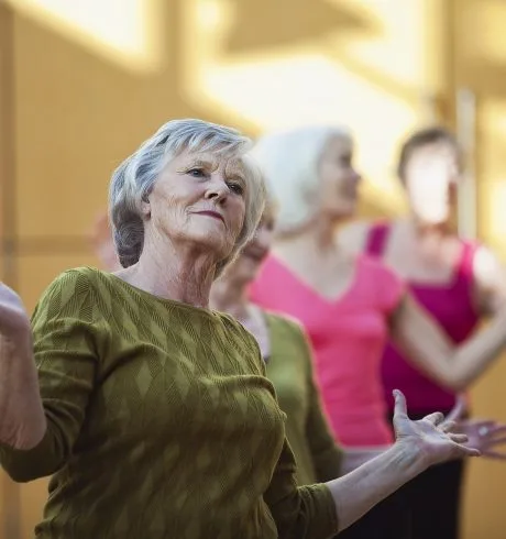 An older woman is performing a dance routine with others. She is leaning back slightly with her arms semi-raised