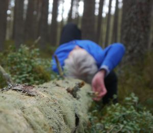 A woman lies on her back on a lichen coverd fallen tree. She has grey hair and is wearing a blue hiking top. SHe is alone in the landscape.