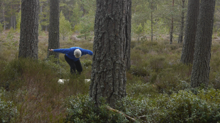 A woman is dancing in a Scottish forest, she is leaning forward with her arms stretched like wings