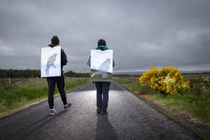Two people walk along an isolated Scottish road. On their backs they have posters featuring pen drawings of birds of prey.