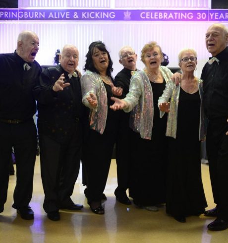 Four older men and three older women are lined up and singing. Their black formal outfits are offset by silver sparkly jackets for the women and silver sequin bow ties for the men.