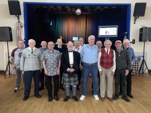 A group of 13 older men stand in a hall in front of a stage. In the centre, one man is in his dress kilt, another wears a red tartan waistcoat