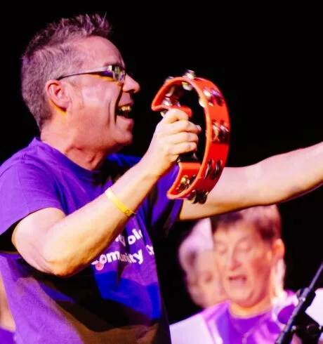 Photo of singing leader Bryan Marshall wearing a purple T-shirt and holding a tambourine with singers in the background
