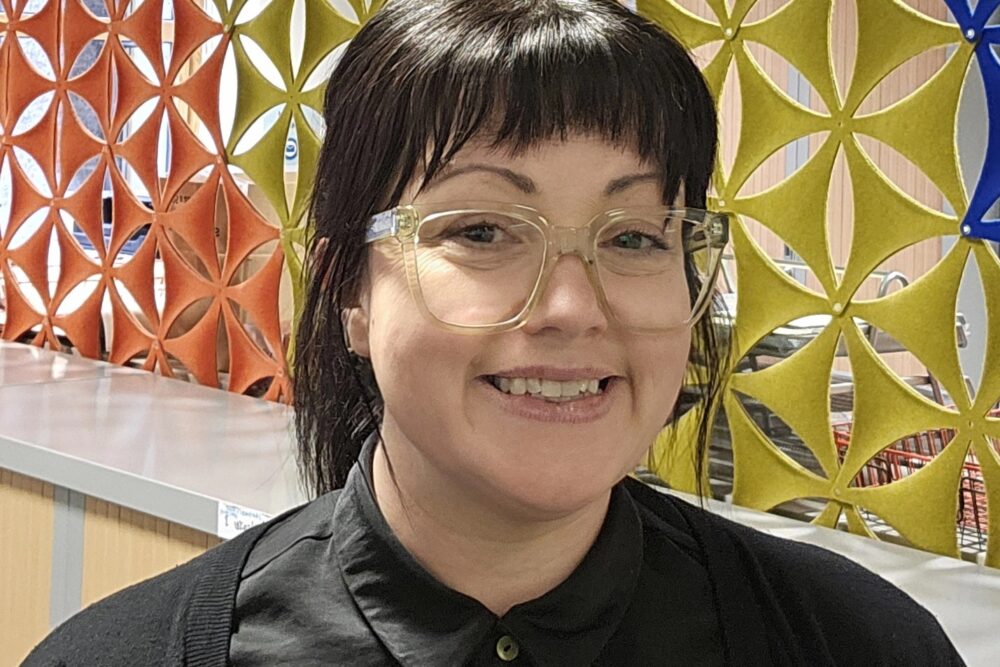 Xana Marwick, Luminate's Administrative Officer, wearing a black shirt and standing in front of a colourful room divider.