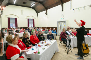 A hall full of people in festive dress sitting at tables and singing with a song leader in Santa hat at the front