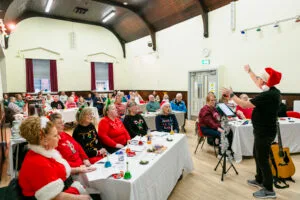 A hall full of people in festive dress sitting at tables and singing with a song leader in Santa hat at the front