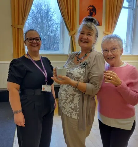Three smiling women standing in a room in front of windows.