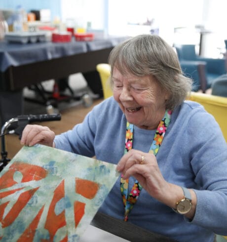 An older woman in a blue sweater smiles and looks at her newly created screenprint