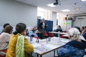 A woman stands in front of tables with art workshop participants in a community centre