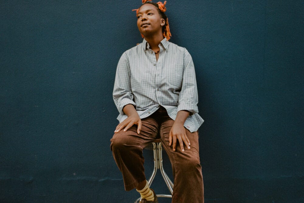 Portrait of artist Jj Fadaka sitting on a stool against a dark blue wall