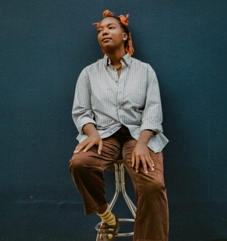 Portrait of artist Jj Fadaka sitting on a stool against a dark blue wall