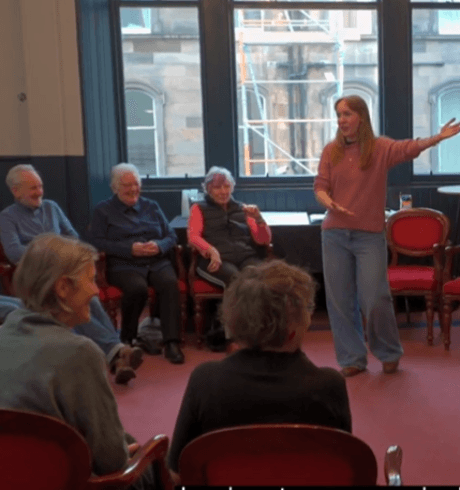 An acting group sit in a circle as the group leader stands up with arm outstretched.