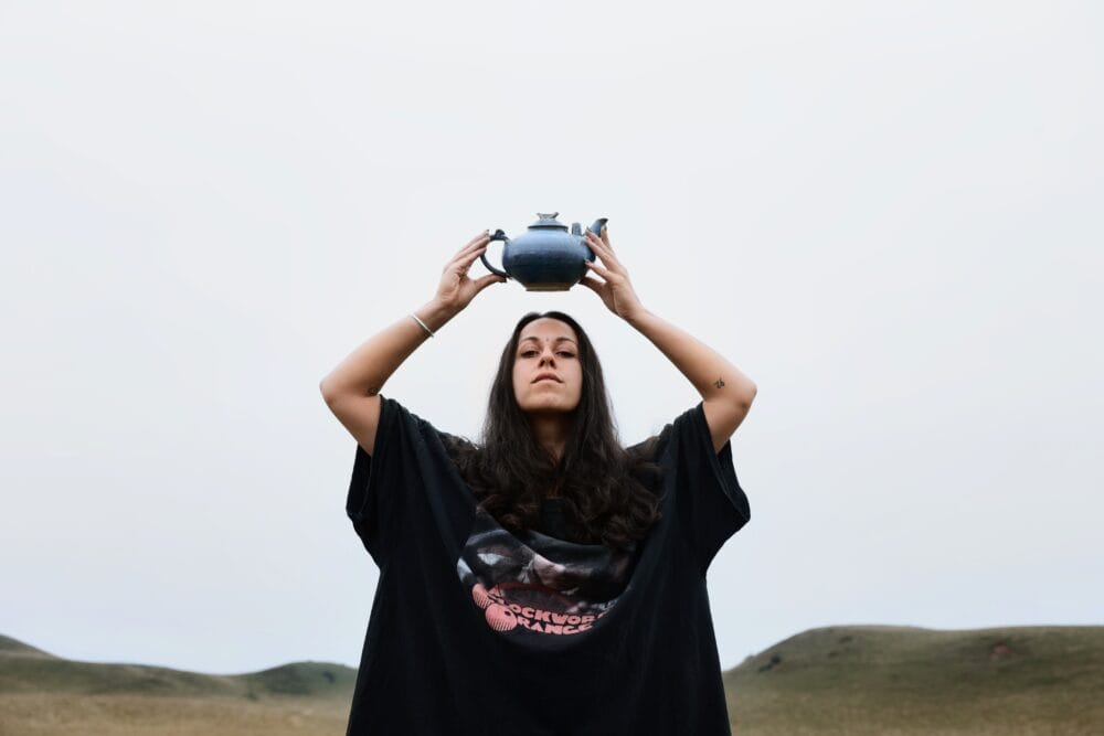 Artist Bee Asha outdoors in a large black T-shirt holding a teapot above her head.