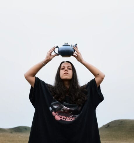 Artist Bee Asha outdoors in a large black T-shirt holding a teapot above her head.