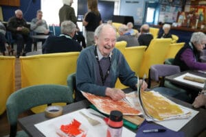 A man laughs while seated with art materials on the table in front of him at Lochside Connections Meeting Centre.