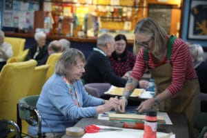 An artist helps a seated woman do screenprinting at Lochside Connections Meeting Centre.