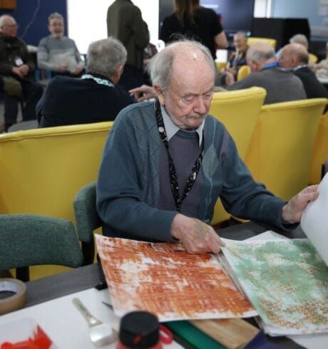 A man sits at a table looking through colourful pieces of paper. A group of people are seated behind him on yellow chairs.