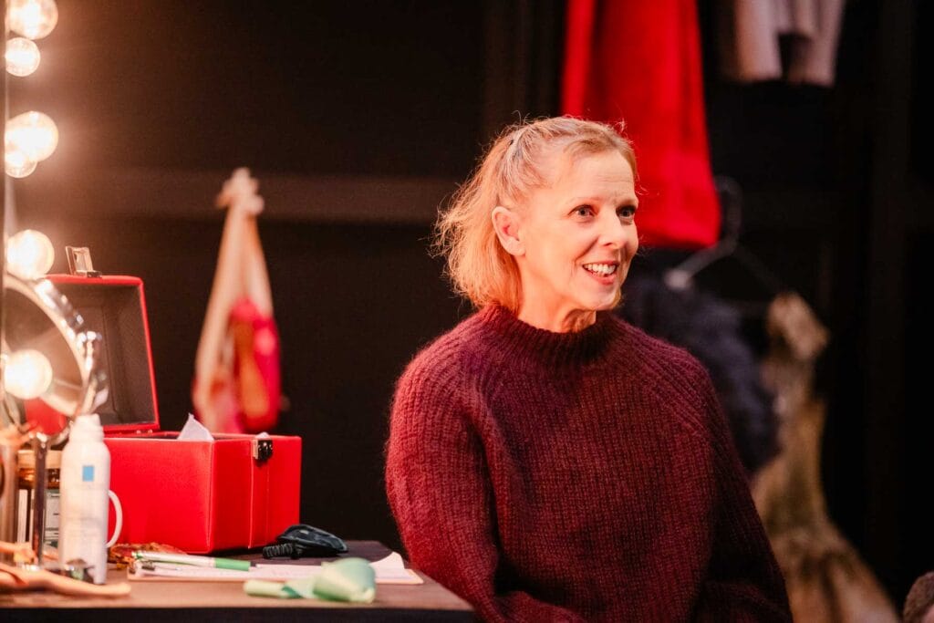 Director and choreographer Kally Lloyd-Jones sits in front of a dressing room mirror wearing a red sweater.