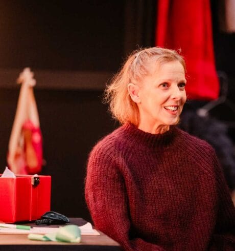 Director and choreographer Kally Lloyd-Jones sits in front of a dressing room mirror wearing a red sweater.